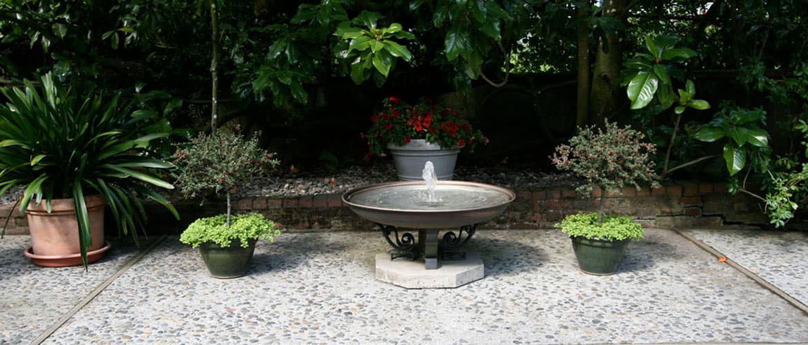 Outdoor patio scene with concrete and river stone floor, potted plants with lush greenery in the background, with a simple bowl-shaped water fountain in the center