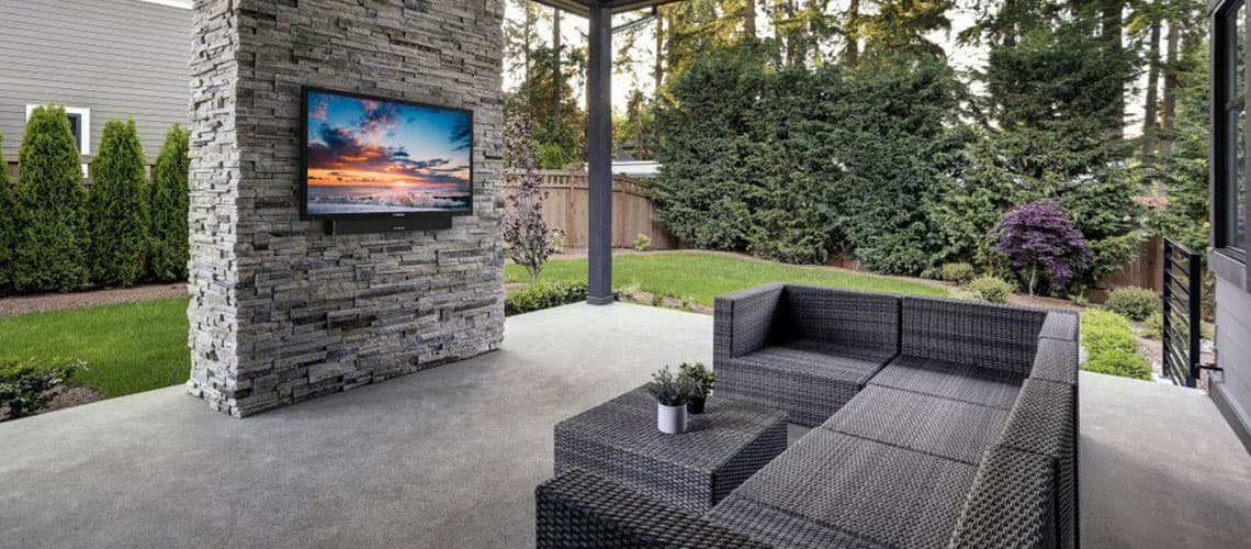 Outdoor residential veranda scene with cement floor, wicker L-shaped couch and coffee table, natural pine slat constructed roof, stacked stone column with a Furrion FDUF55CBR full shade outdoor TV hung on it