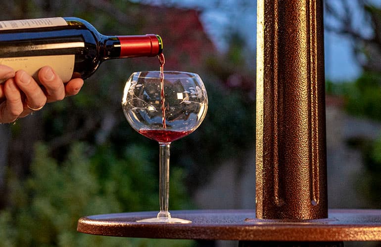 Close up image of bronze table built into patio heater, with wine glass upon it being filled by man pouring red wine with a blurred outdoor background.