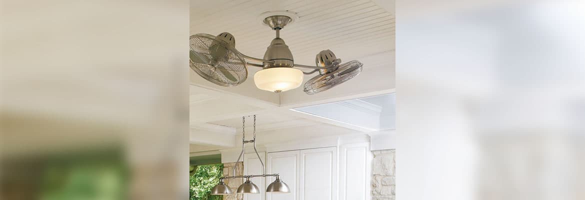 Outdoor kitchen scene with mortared stone walls, white cabinets, stainless-steel grill, gray countertops, gray iron barstools with beige cushions, food set on the countertops, white coffered ceiling from which hangs a three-light trestle fixture and a Craftmade Lighting Bellows II BW248BNK6 dual head ceiling fan