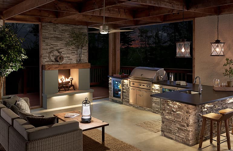 Outdoor kitchen at sunset, large stacked white stone counter with built-in grill with Kichler 49744 pendant lights in weathered zinc, couch seating area with rustic coffee table, open fireplace with stacked stone, and Kichler 310155NI ceiling fan on walnut colored thin beam ceiling.