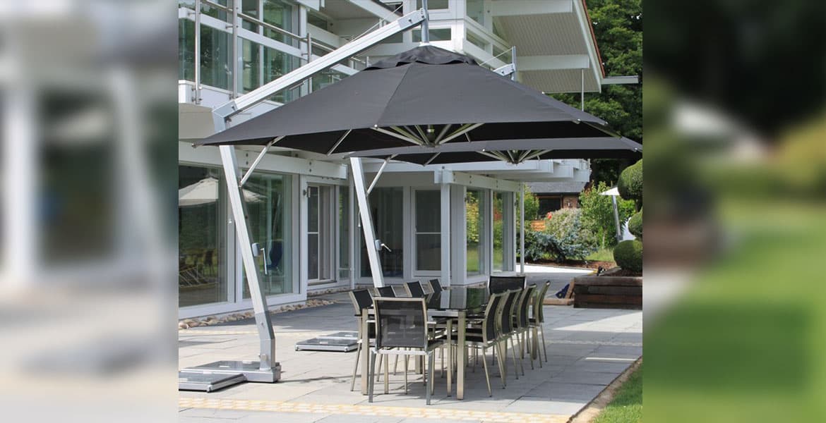 Outdoor patio scene in commercial building area with concrete patio , lush green lawns, large metal and glass building in the background with a large outdoor dining area with metal framed chairs and table, black seating in the chairs and smoked glass top on the table, over which looms the shade from a Bambrella Hurricane Side Wind 3-4MSQ-SW-H aluminum cantilever umbrella